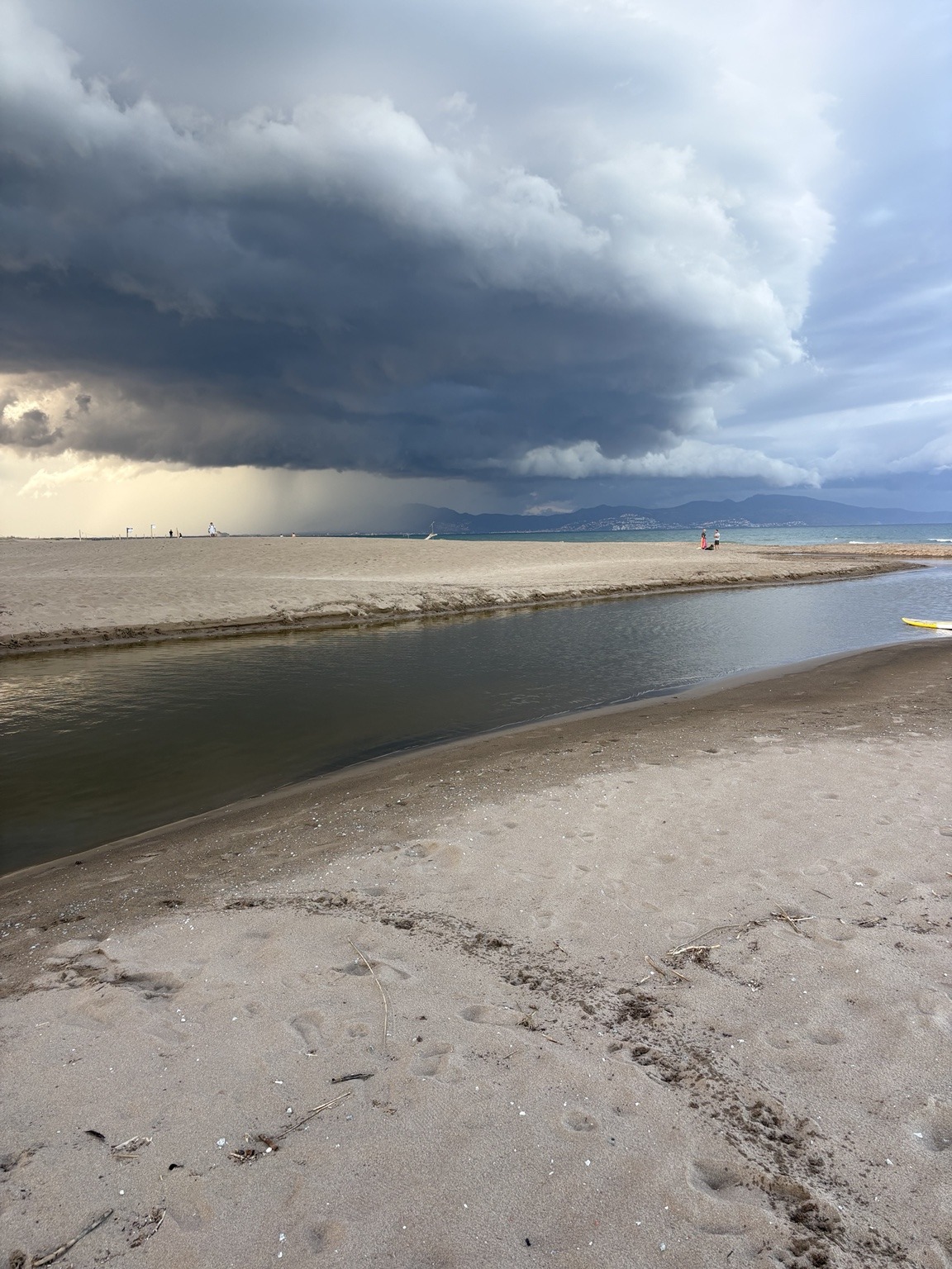 The broad Costa Brava shoreline under storm clouds, a shallow stream crossing the sand and mountains visible across the bay.