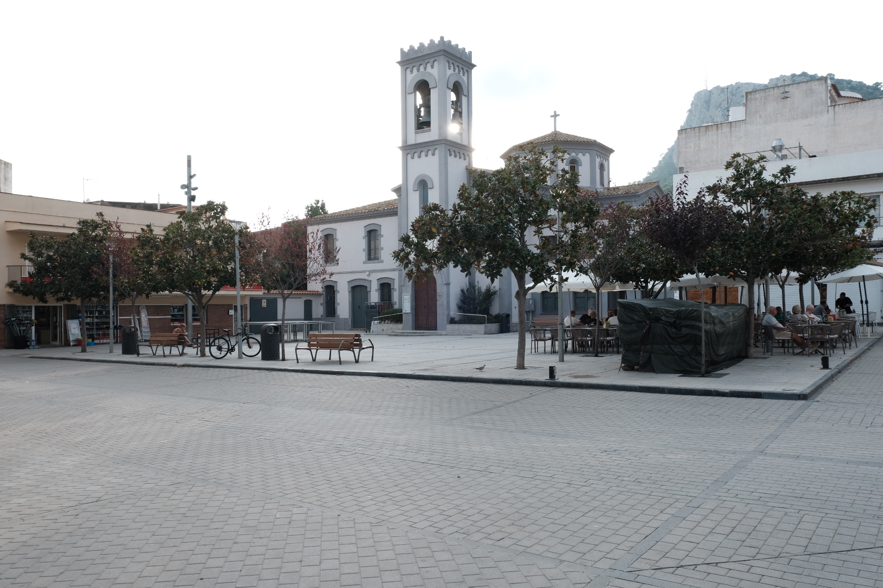 A paved village square with benches, trees and café tables in front of a white church and bell tower.