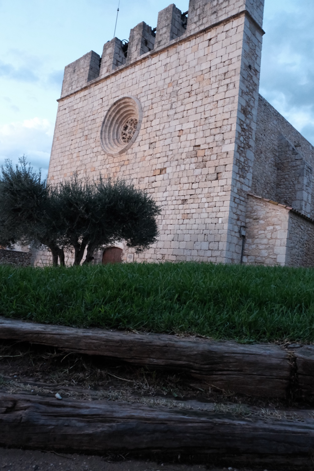A stone church with a circular window and bell openings, framed by olive trees, grass and a wooden fence.