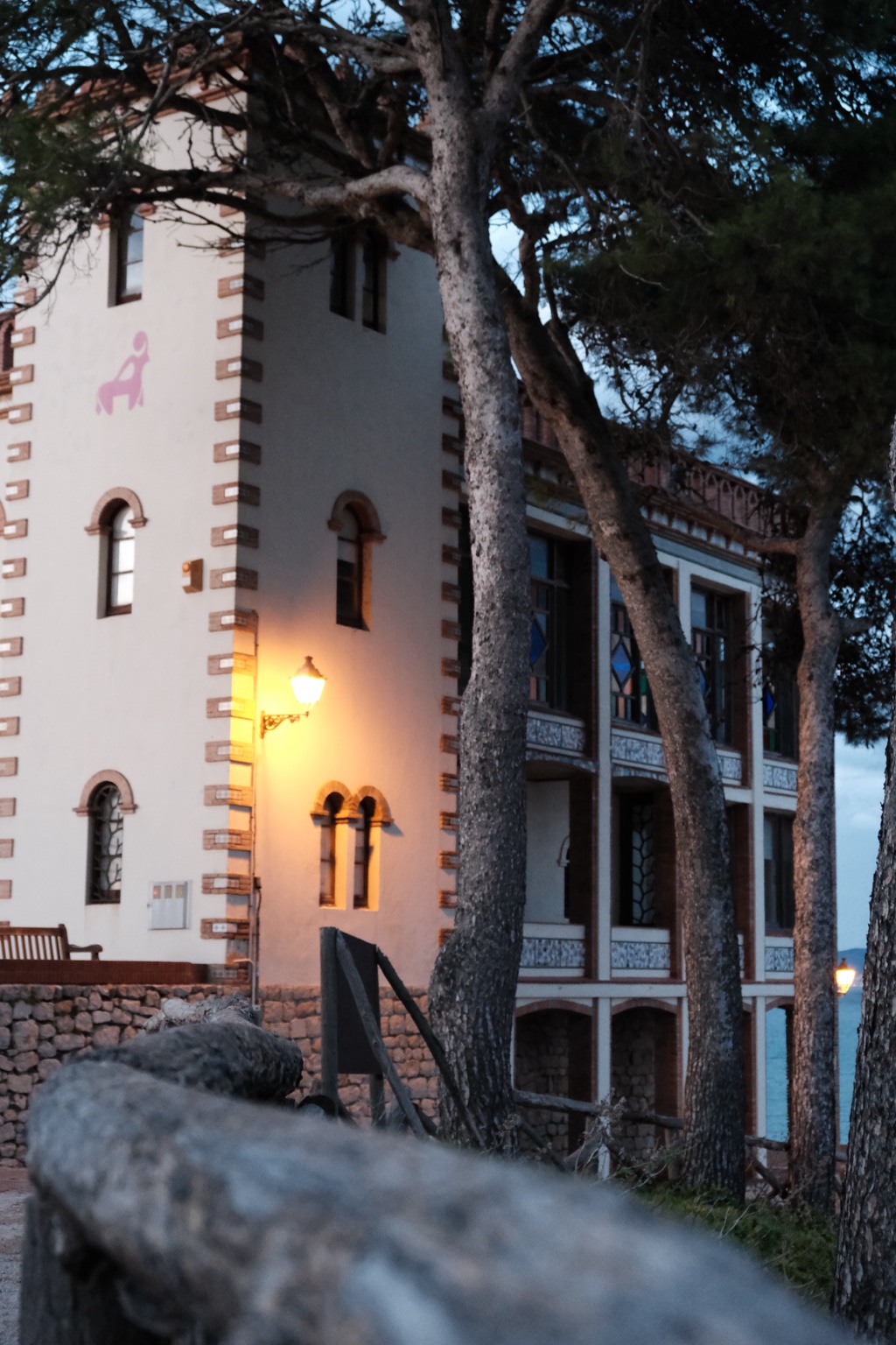 A pale building with arched windows and balconies among pine trees, lit by a warm exterior lamp at dusk.