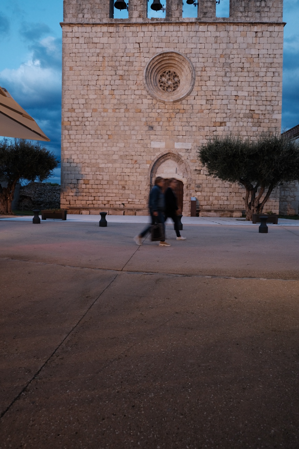 Two people walk across a square in front of a stone church façade with a circular window and bells.