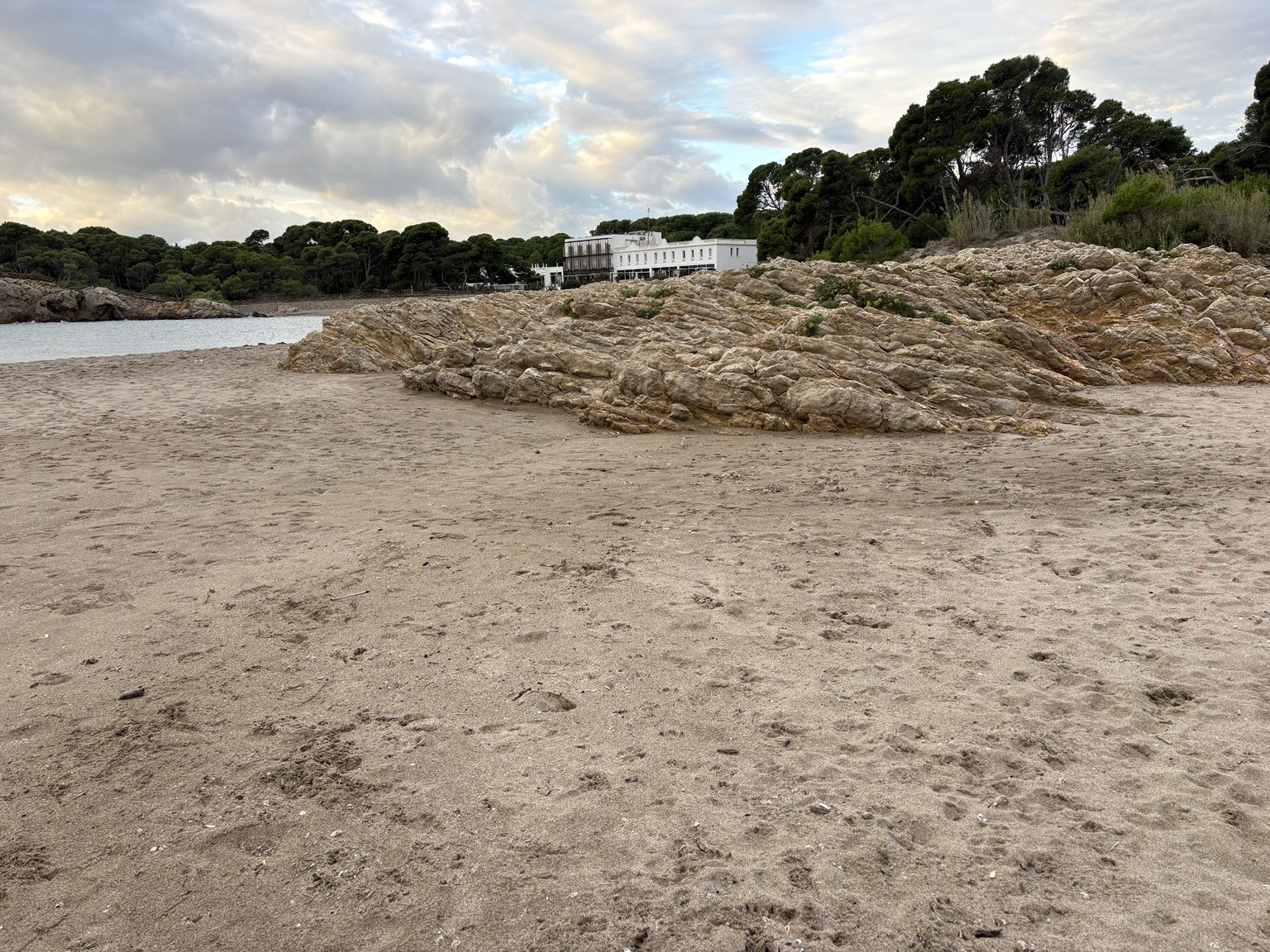 A wide, quiet view across the sandy beach toward a rocky shoreline, with pines and a white building in the distance under soft evening clouds.