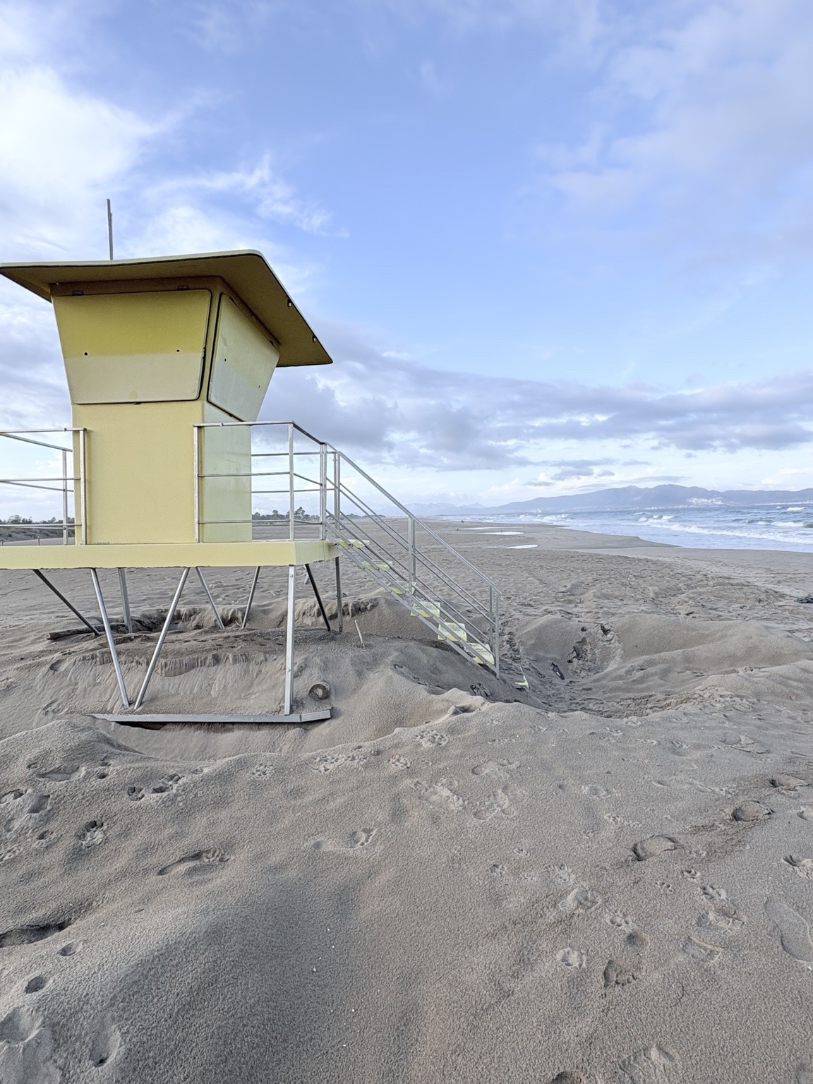 A pale yellow lifeguard hut stands above an eroded sandy beach, with the sea and distant coastline beyond.