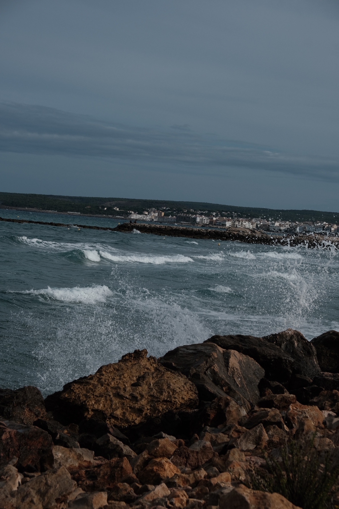 Waves break against foreground rocks, with a coastal town across the bay under a cloudy sky.