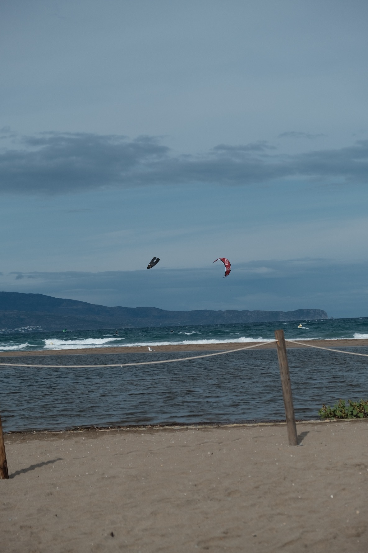 Two kitesurfing kites — one black, one red — above the water, with the distant coast and mountains beyond.