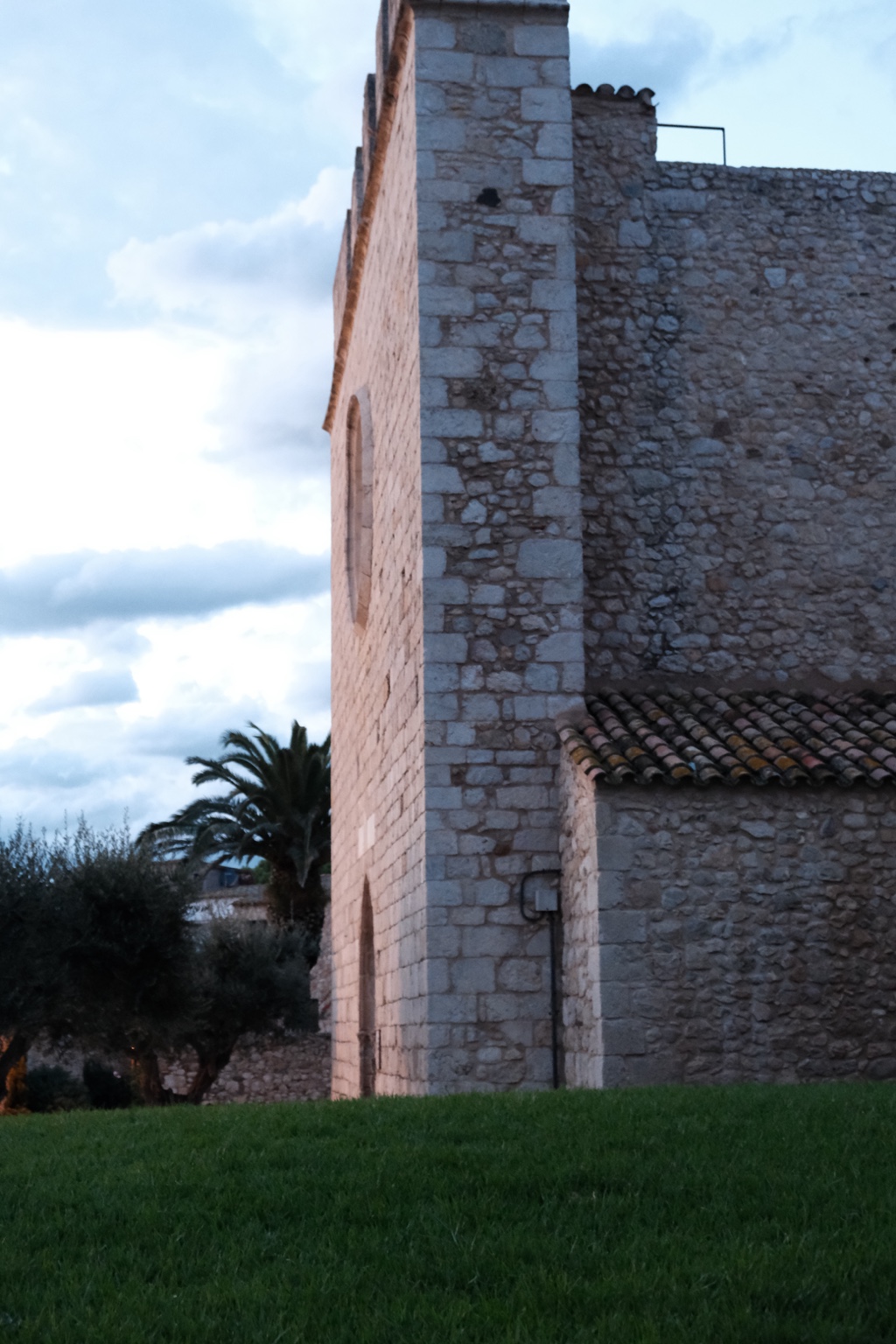 A close view of a stone church corner and tiled roof above grass, with palms and olive trees beyond.